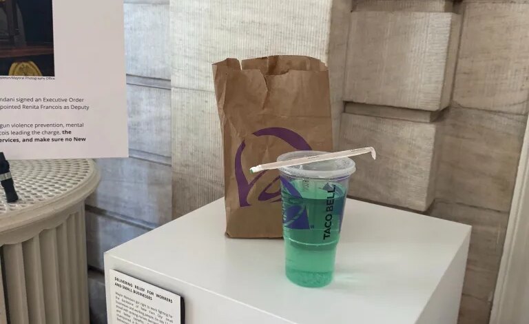 A Taco Bell Baja Blast drink with a straw, a brown Taco Bell paper bag, and a plaque about "Delivering Relief for Workers and Small Businesses" on a white pedestal in what appears to be a museum exhibit.