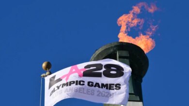 A white LA28 Olympic Games flag waving in front of a lit Olympic cauldron against a clear blue sky.