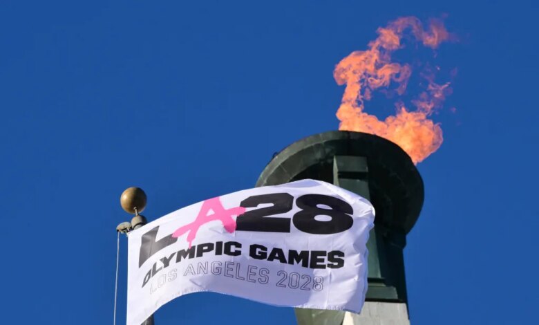 A white LA28 Olympic Games flag waving in front of a lit Olympic cauldron against a clear blue sky.