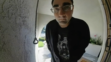 A man in glasses, a black sweatshirt, and dark pants, stands on a "Welcome" mat, looking directly into a Ring doorbell camera.