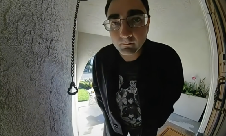 A man in glasses, a black sweatshirt, and dark pants, stands on a "Welcome" mat, looking directly into a Ring doorbell camera.