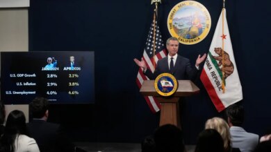 Governor Gavin Newsom speaks at a podium next to a screen displaying US economic statistics and images of Joe Biden and Donald Trump.