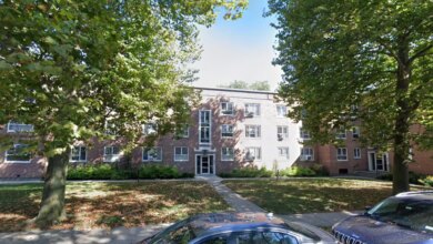 A multi-story brick apartment building with a sidewalk leading to the front door, surrounded by trees and a grassy lawn with fallen leaves, with two cars parked in the foreground.