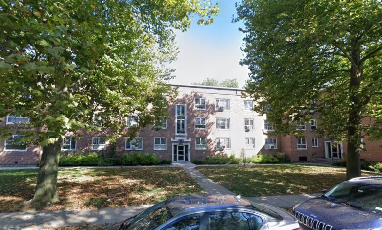 A multi-story brick apartment building with a sidewalk leading to the front door, surrounded by trees and a grassy lawn with fallen leaves, with two cars parked in the foreground.
