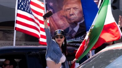 Woman leaning out of a car in a procession, holding up her phone while an American flag and a banner with Donald Trump's face are visible in the background.