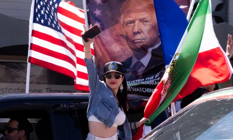 Woman leaning out of a car in a procession, holding up her phone while an American flag and a banner with Donald Trump's face are visible in the background.