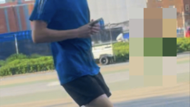 A cropped image of an individual sought by the NYPD, wearing a blue shirt and black shorts, walking on a street.