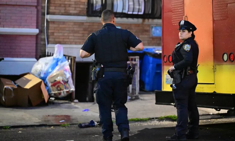 Two police officers stand on a street, with one facing away and the other looking towards the right, near a building and a yellow vehicle.