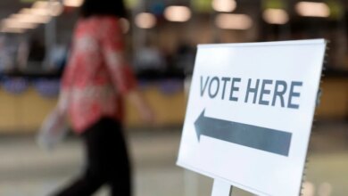 A "VOTE HERE" sign with an arrow points left while a person walks past in the background.