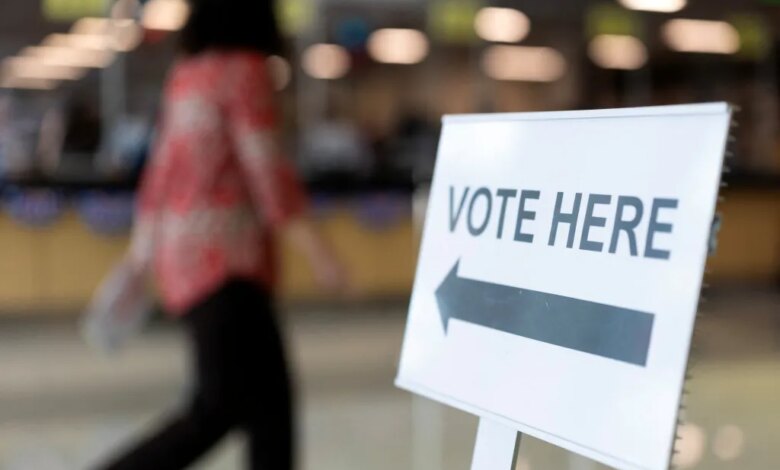 A "VOTE HERE" sign with an arrow points left while a person walks past in the background.