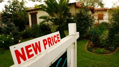 A "NEW PRICE" real estate sign in front of a stucco house with a tile roof and green landscaping.