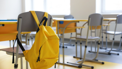 A bright yellow backpack hangs on the back of a grey school chair in a classroom with yellow desks and additional chairs.