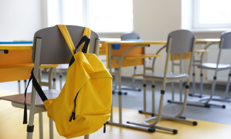 A bright yellow backpack hangs on the back of a grey school chair in a classroom with yellow desks and additional chairs.