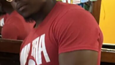 Still image from surveillance video of a dark-skinned male with gold-rimmed circular sunglasses and a red t-shirt with white text, suspected of sexual assault.