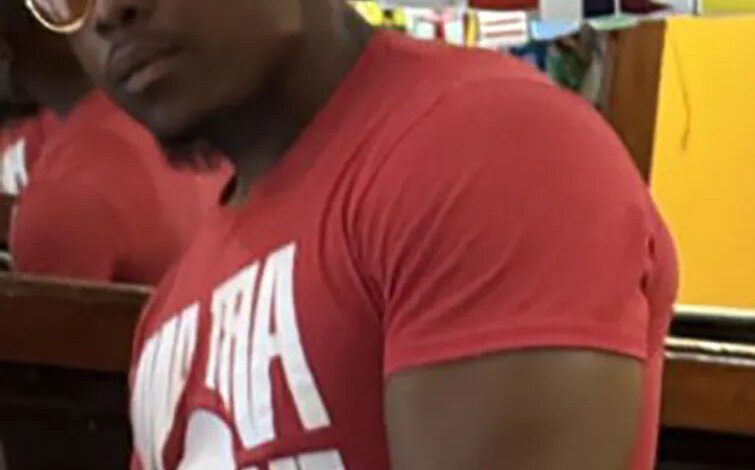 Still image from surveillance video of a dark-skinned male with gold-rimmed circular sunglasses and a red t-shirt with white text, suspected of sexual assault.