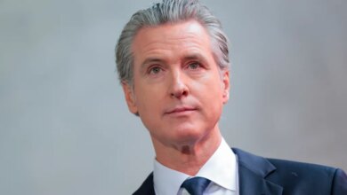 California Governor Gavin Newsom, dressed in a dark suit and blue tie, looks on.