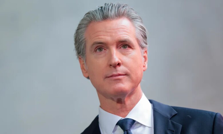 California Governor Gavin Newsom, dressed in a dark suit and blue tie, looks on.
