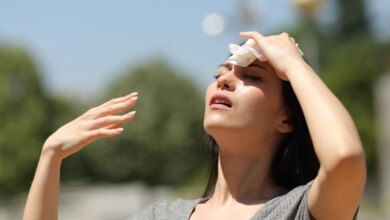 A young Asian woman wipes sweat from her forehead with a tissue while looking up, fanning herself with her other hand, in a park on a hot summer day.