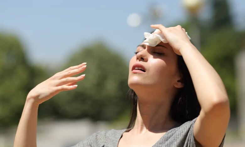A young Asian woman wipes sweat from her forehead with a tissue while looking up, fanning herself with her other hand, in a park on a hot summer day.