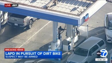 Aerial view of a person wearing a black hoodie and mask, pumping gas at a Chevron station, with cars around the pumps, as "LAPD IN PURSUIT OF DIRT BIKE SUSPECT MAY BE ARMED" appears on screen.