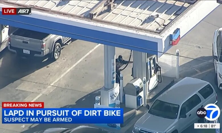 Aerial view of a person wearing a black hoodie and mask, pumping gas at a Chevron station, with cars around the pumps, as "LAPD IN PURSUIT OF DIRT BIKE SUSPECT MAY BE ARMED" appears on screen.