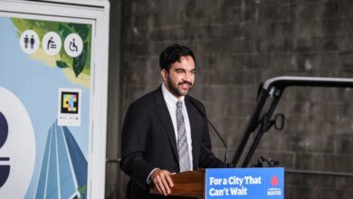 NYC Mayor Zohran Mamdani speaking at a podium with "For a City That Can't Wait" sign, next to a sign with public restroom symbols.