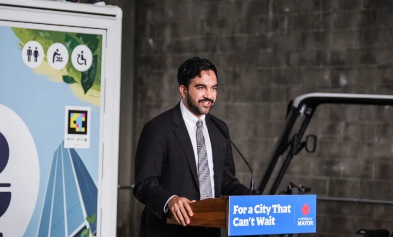 NYC Mayor Zohran Mamdani speaking at a podium with "For a City That Can't Wait" sign, next to a sign with public restroom symbols.