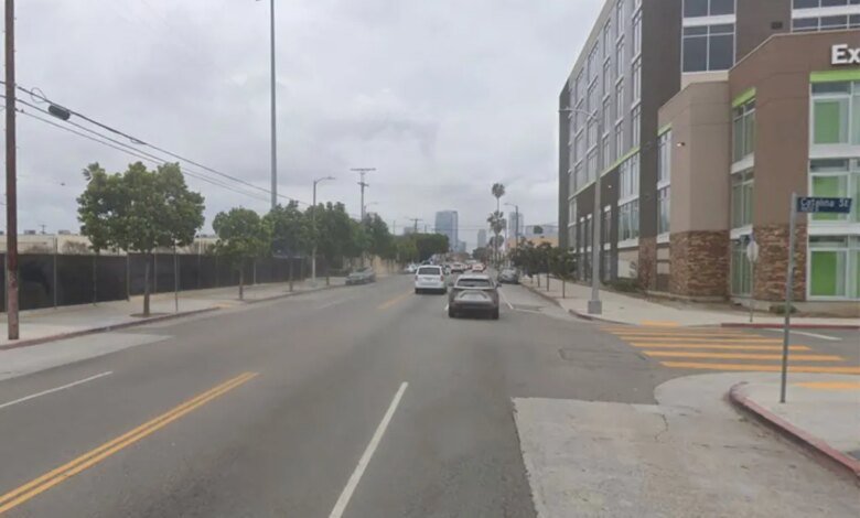 Street level view of a city street under an overcast sky with modern buildings and trees lining the road, and cars driving away in the distance.